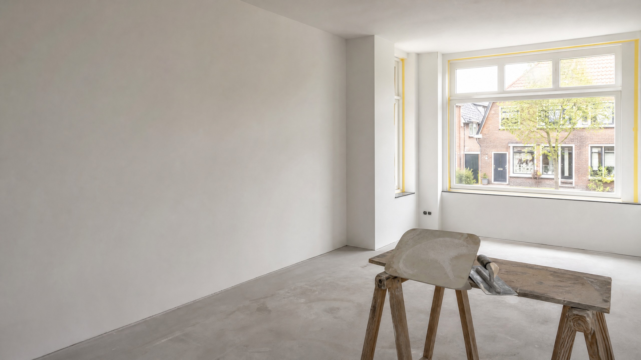 Sfeerbeeld: vers gestucte woonkamer met gladde witte wanden, een raam met gele afplaktape en een spaan op een schraag